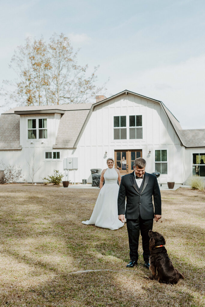 A bride in a white dress stands outside a white house, watching a man in a suit and bow tie walking a brown dog on a lawn during their first look at The Venues at Ogeechee Tech