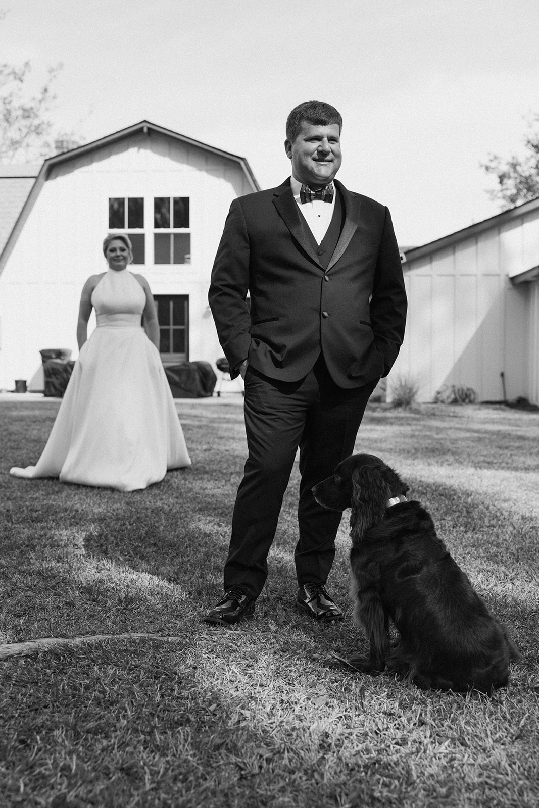 A woman in a white dress smiles at and pets a dark-colored dog with a red collar, outside in front of a white buildings at The Venues at Ogeechee Tech
