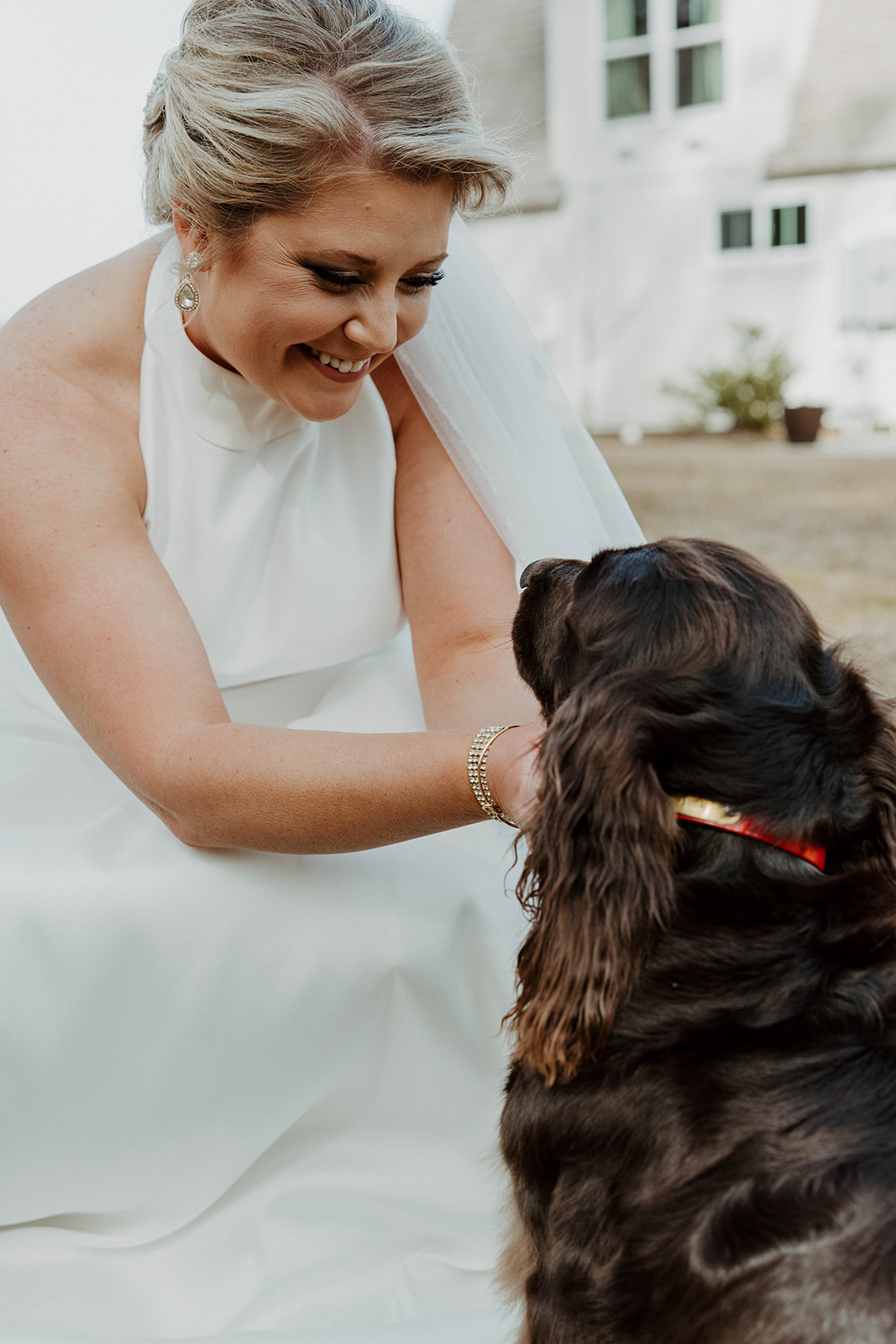 A woman in a white dress smiles at and pets a dark-colored dog with a red collar, outside in front of a white buildings at The Venues at Ogeechee Tech