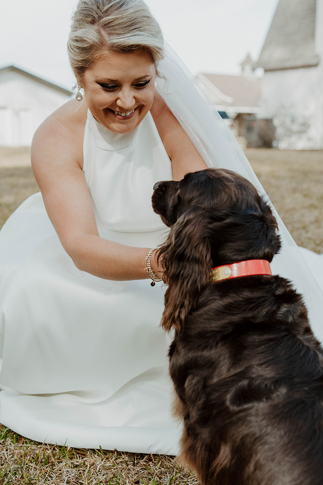 A woman in a white dress smiles at and pets a dark-colored dog with a red collar, outside in front of a white buildings at The Venues at Ogeechee Tech