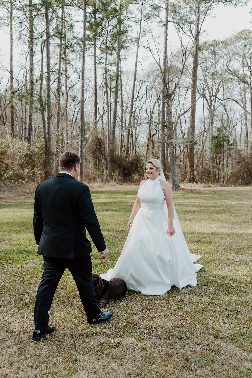 A bride in a white dress stands outside a white house, watching a man in a suit and bow tie walking a brown dog on a lawn during their first look at The Venues at Ogeechee Tech