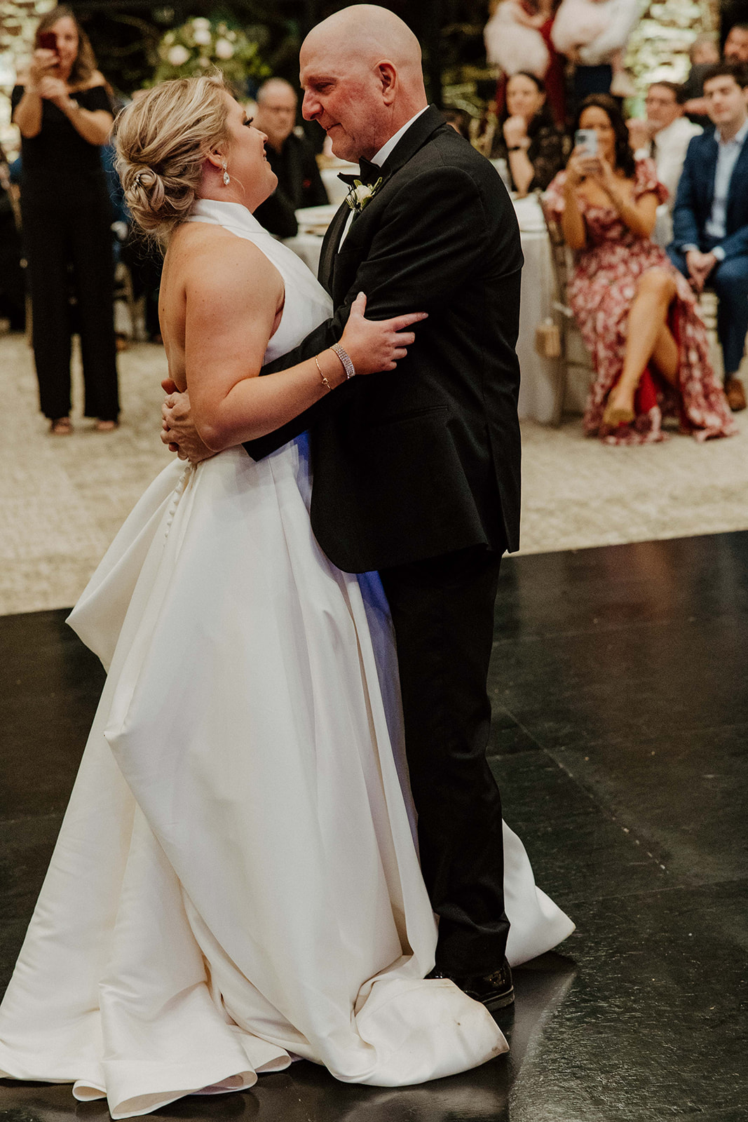 A couple dances on a black floor in a large, well-lit room with stone columns and seated guests watching them in the background. The woman is wearing a white gown, and the man is in a suit.