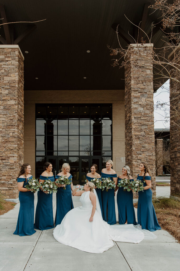 A bride in a white gown stands in front of a building with five bridesmaids dressed in blue, posing with bouquets and smiling.