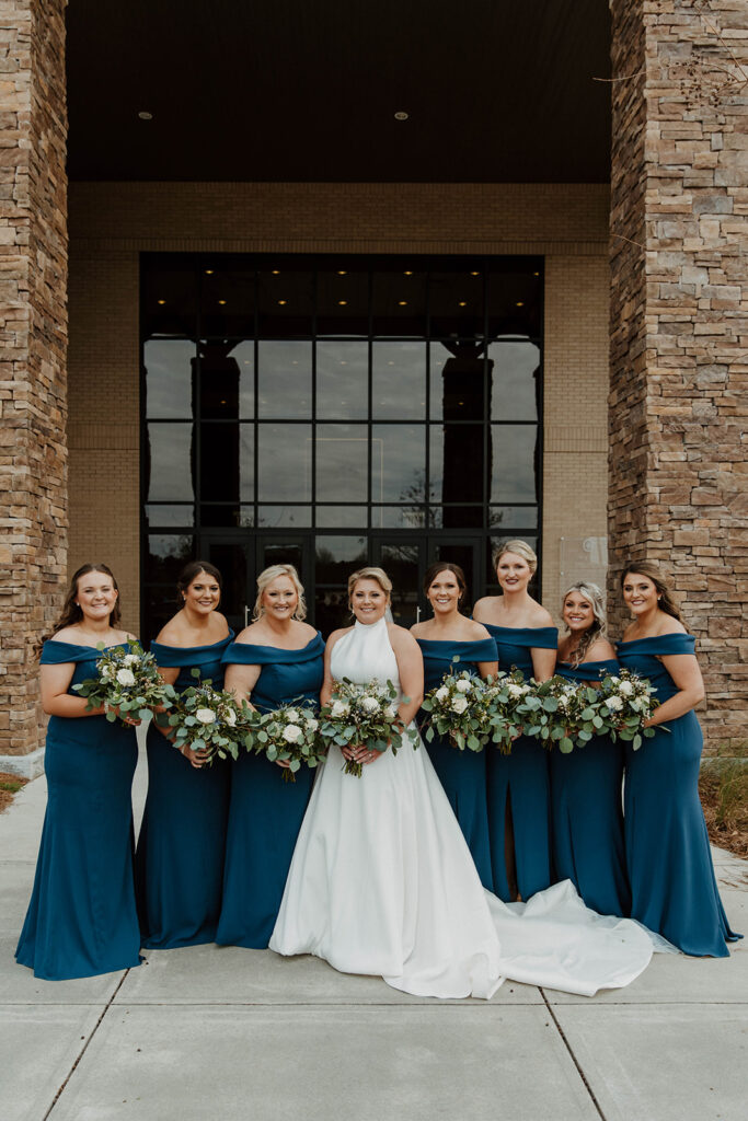 A bride in a white gown stands in front of a building with five bridesmaids dressed in blue, posing with bouquets and smiling.