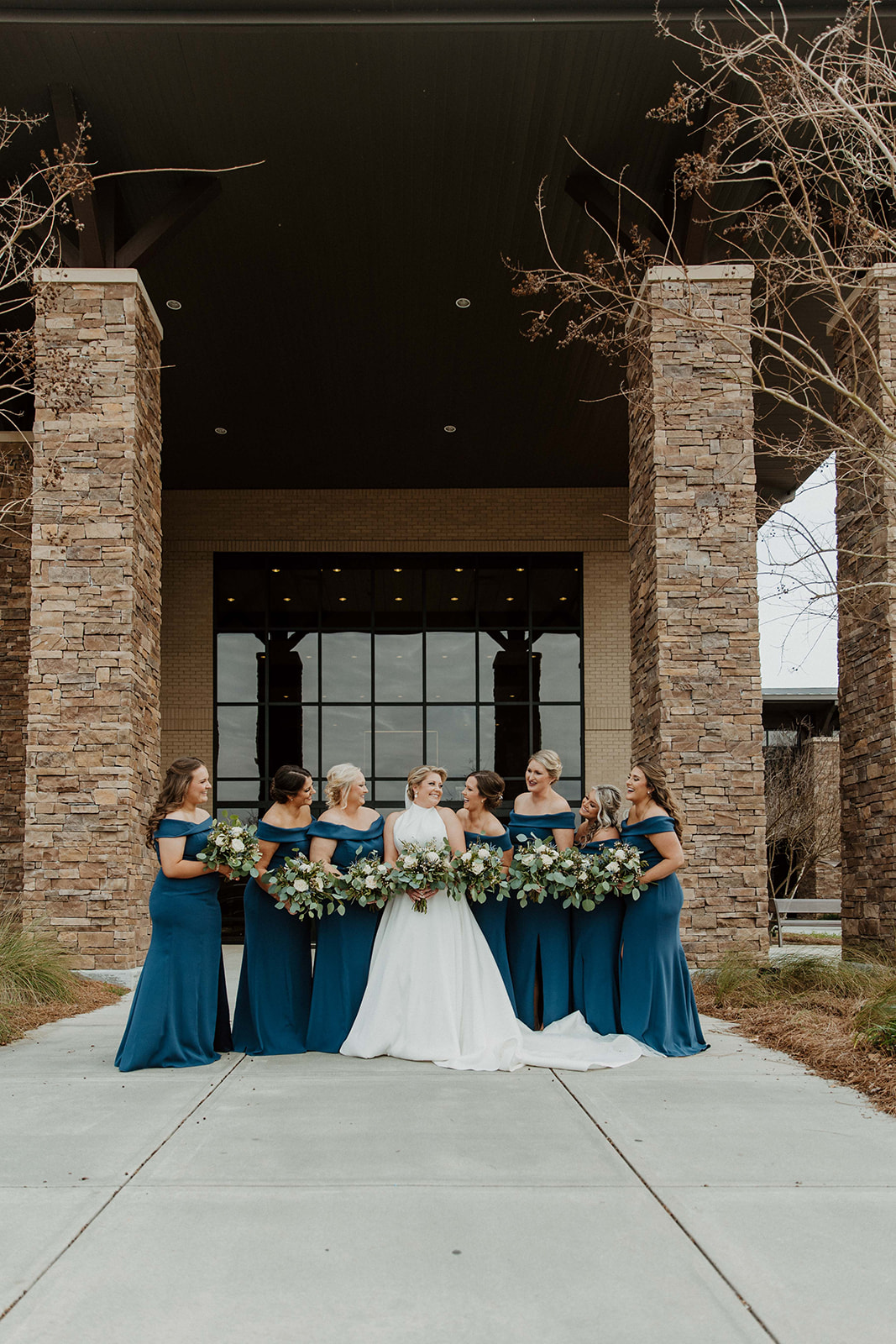 A bride in a white gown stands in front of a building with five bridesmaids dressed in blue, posing with bouquets and smiling.