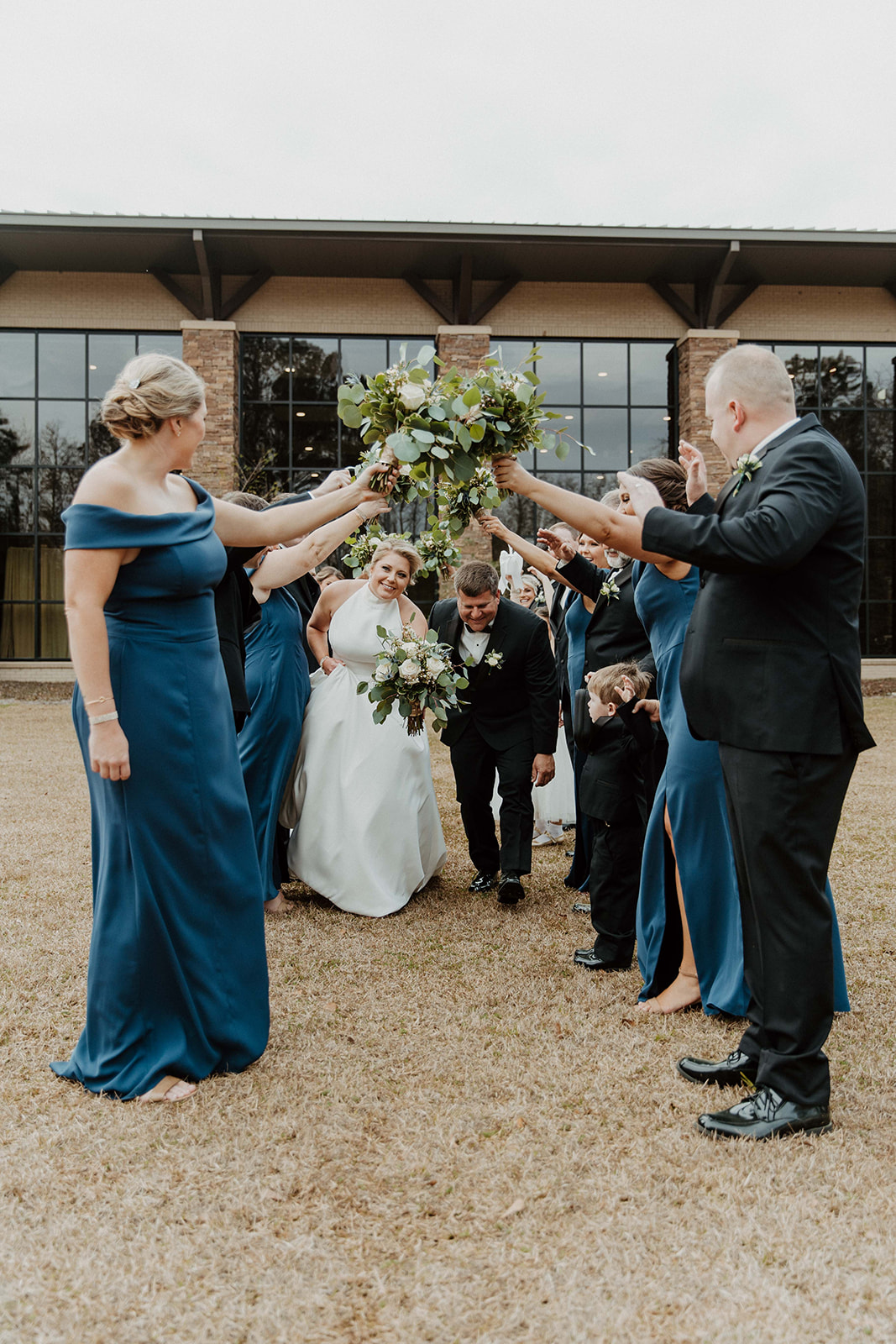 A newlywed couple walks under a tunnel of raised hands holding bouquets, with bridesmaids in blue dresses and groomsmen in dark suits standing on either side.