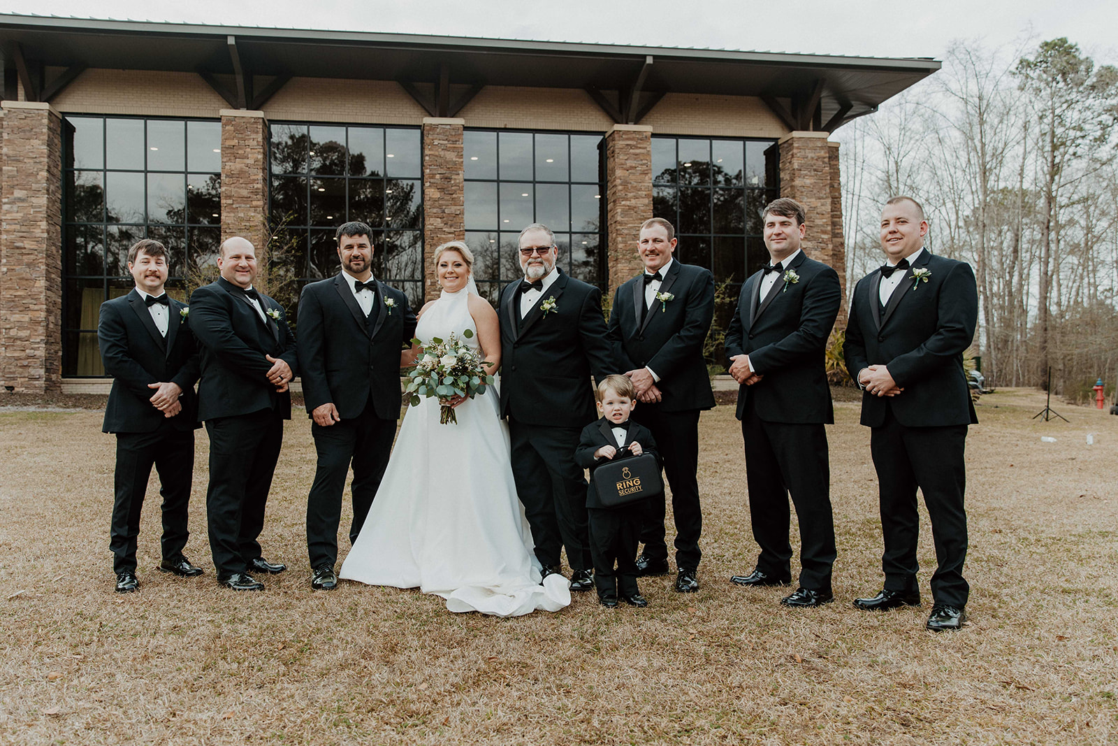 A bride and groom stand with a group of eight men in formal attire, including a young boy in a suit, posing for a photo outside a brick building with large windows.