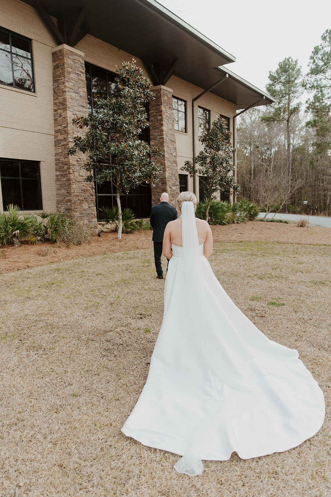 A bride in a white gown with a long train walks towards a man in a suit outside a large building with stone accents and tall windows during a first look with dad at The Venues at Ogeechee Tech
