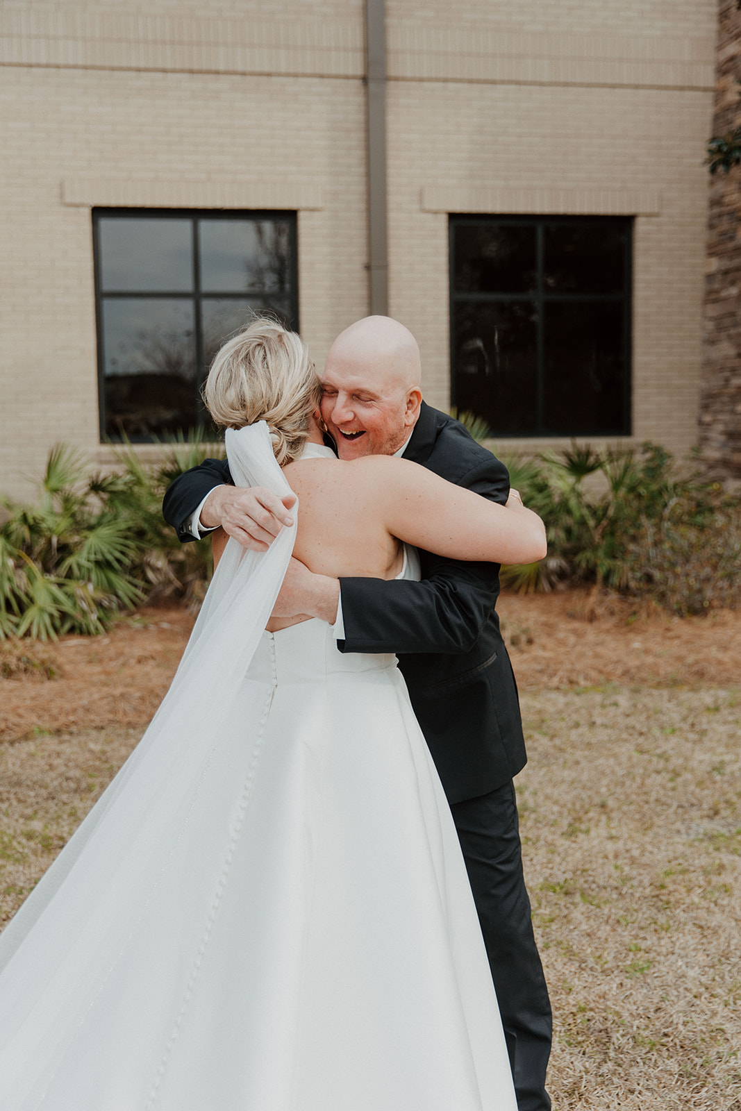 A bride in a white gown with a long train walks towards a man in a suit outside a large building with stone accents and tall windows during a first look with dad at The Venues at Ogeechee Tech