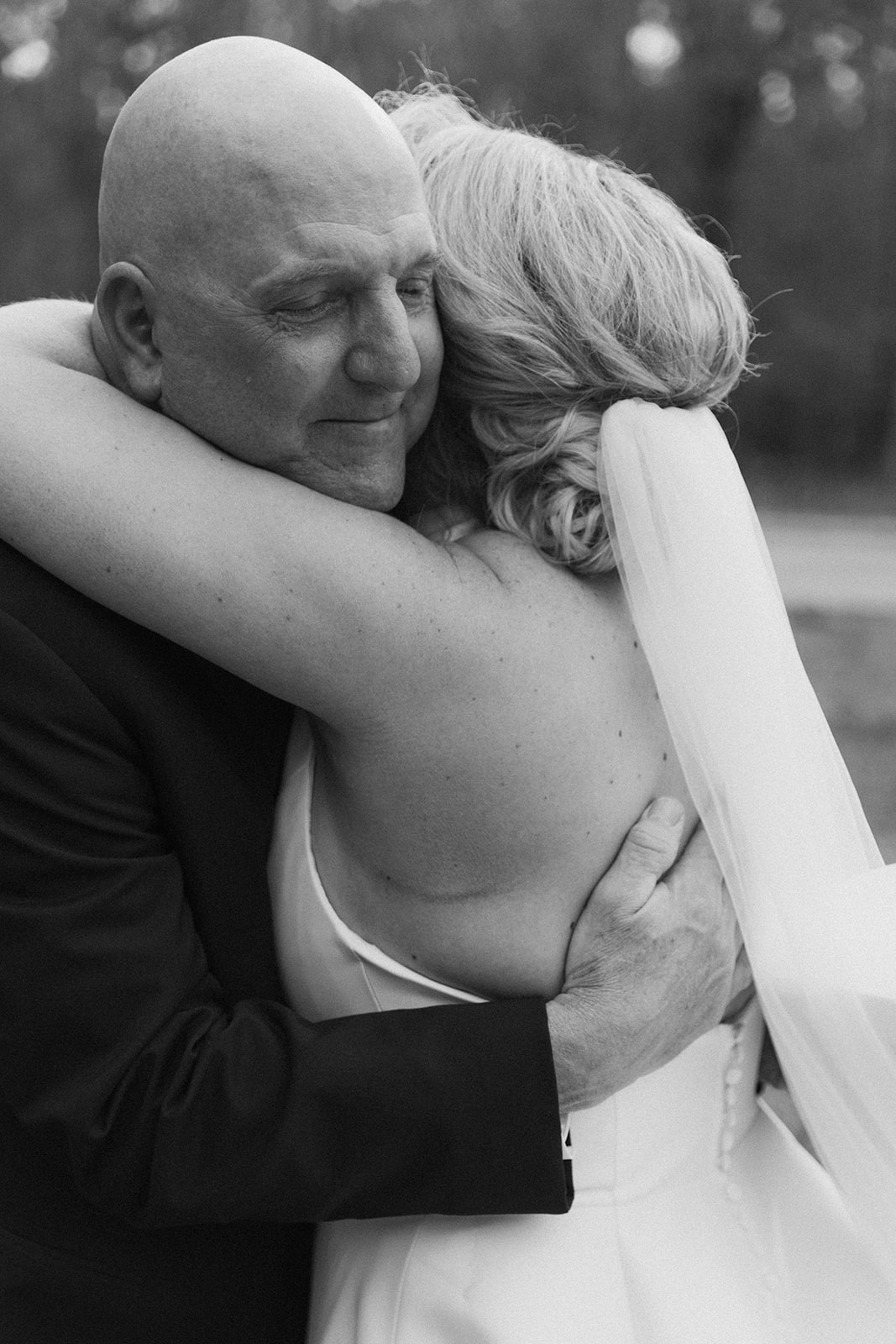 A bride in a white gown with a long train walks towards a man in a suit outside a large building with stone accents and tall windows during a first look with dad at The Venues at Ogeechee Tech