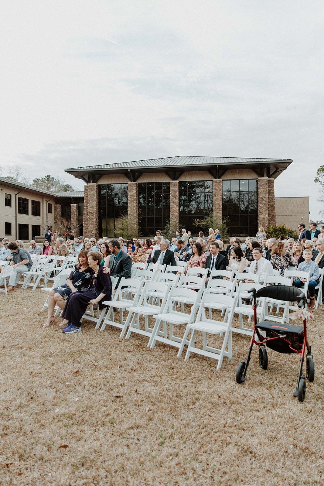 A crowd of people sits on white folding chairs set up on a lawn in front of a modern building with large windows at The Venues at Ogeechee Tech
