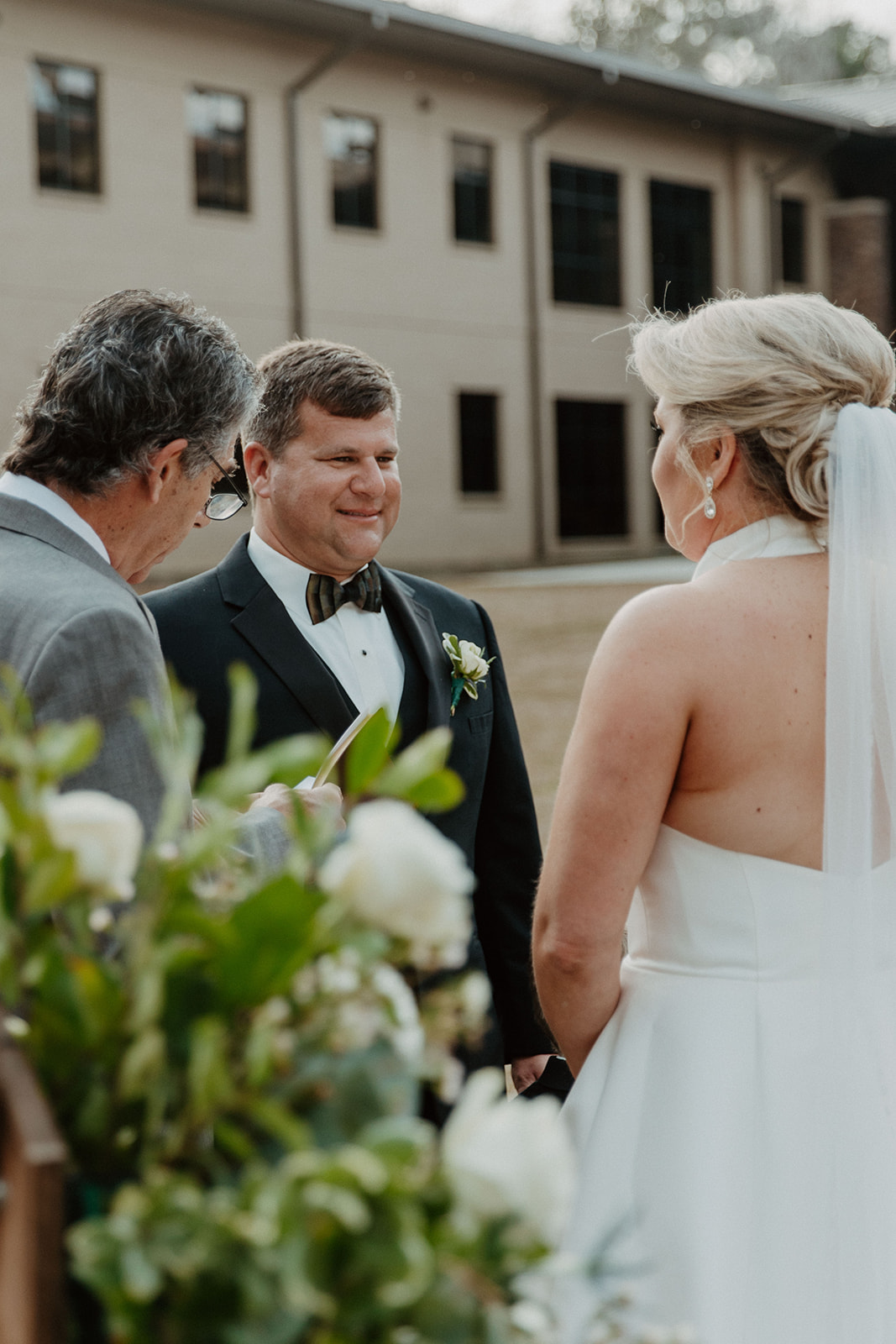 A bride and groom stand facing an officiant during their outdoor wedding ceremony, with a building visible in the background. Flowers are in the foreground at  The Venues at Ogeechee Tech