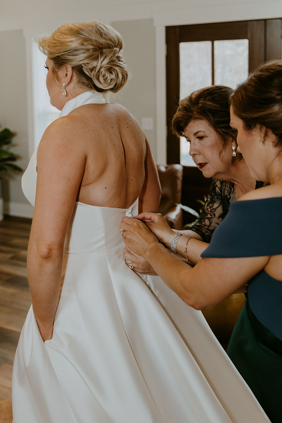 A bride is being assisted by two women as they adjust the back of her white wedding dress. They are indoors, and the room has wood flooring and natural light from the windows at The Venues at Ogeechee Tech
