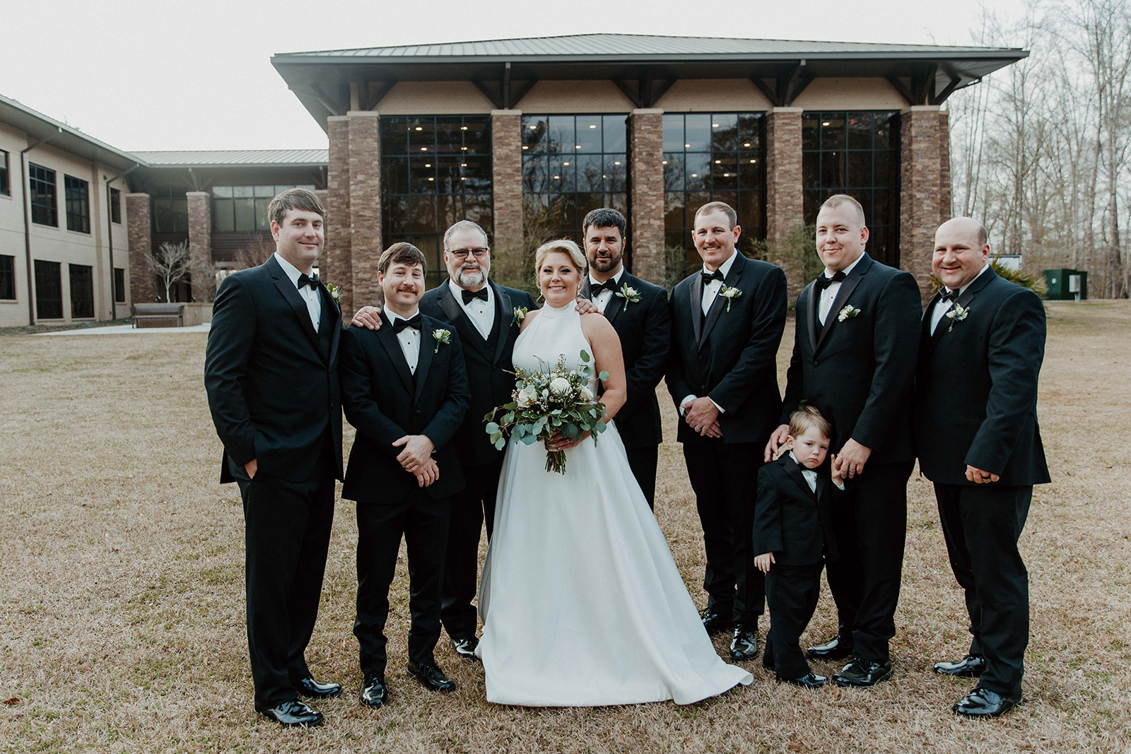 A bride and groom stand with a group of eight men in formal attire, including a young boy in a suit, posing for a photo outside a brick building with large windows.