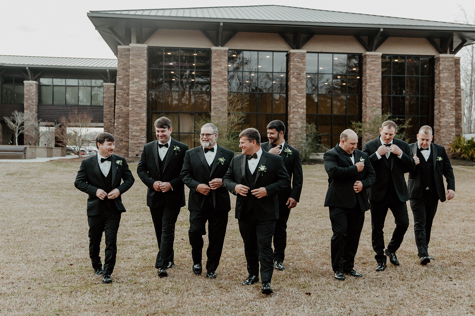 A bride and groom stand with a group of eight men in formal attire, including a young boy in a suit, posing for a photo outside a brick building with large windows.