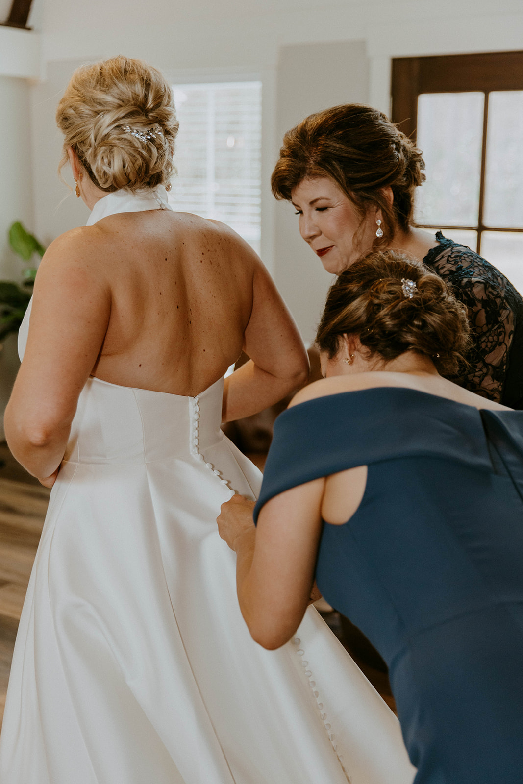 A bride is being assisted by two women as they adjust the back of her white wedding dress. They are indoors, and the room has wood flooring and natural light from the windows at The Venues at Ogeechee Tech