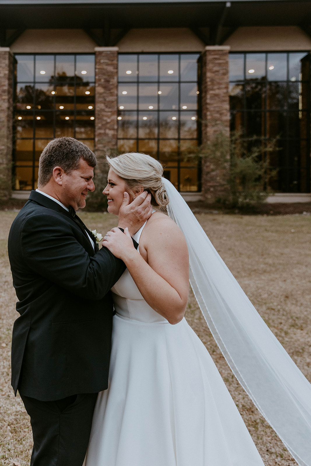 A bride and groom embrace outside a building with large windows. The bride's veil is flowing, and they both have their eyes closed. Trees and foliage are in the background at The Venues at Ogeechee Tech
