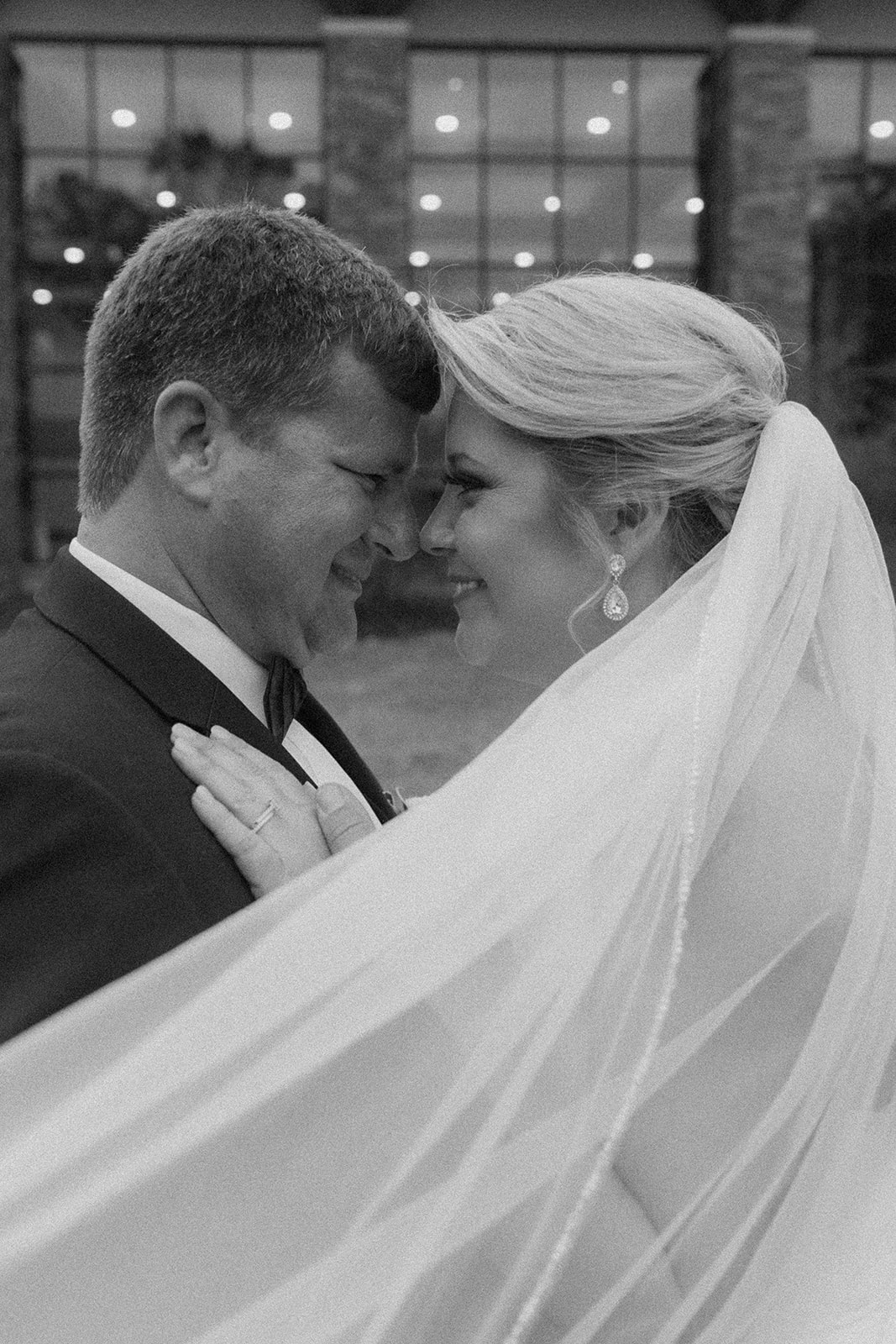 A bride and groom embrace outside a building with large windows. The bride's veil is flowing, and they both have their eyes closed. Trees and foliage are in the background at The Venues at Ogeechee Tech
