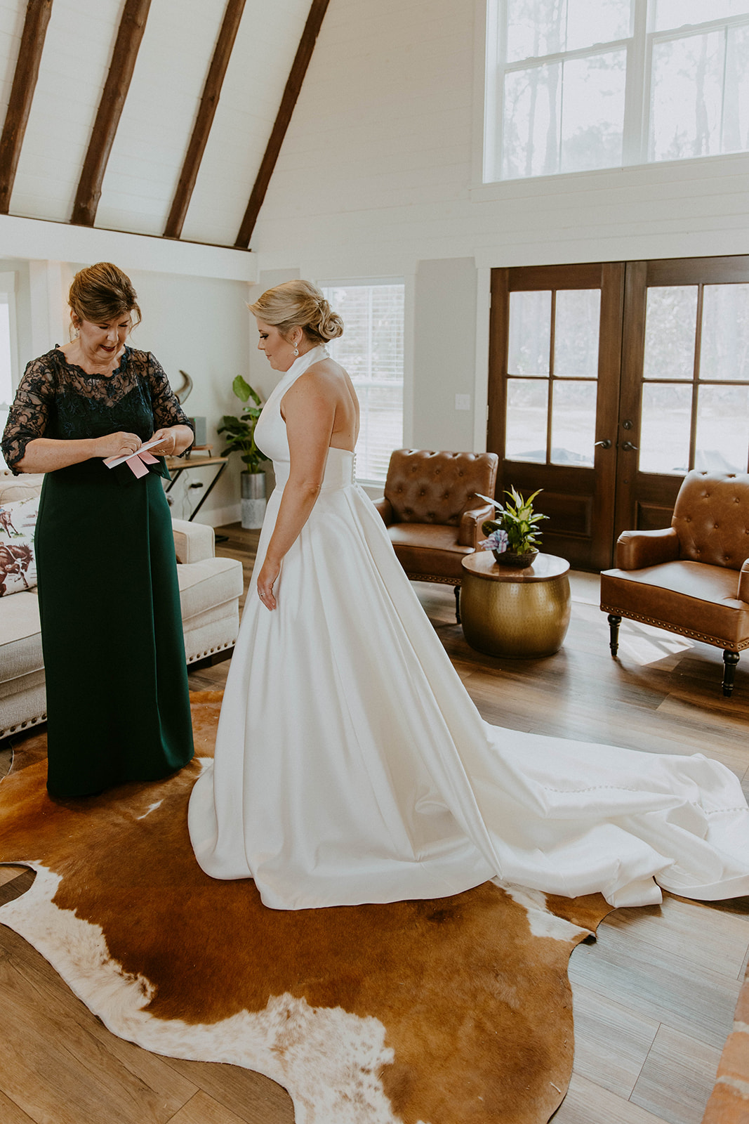 A bride is being assisted by two women as they adjust the back of her white wedding dress. They are indoors, and the room has wood flooring and natural light from the windows at The Venues at Ogeechee Tech