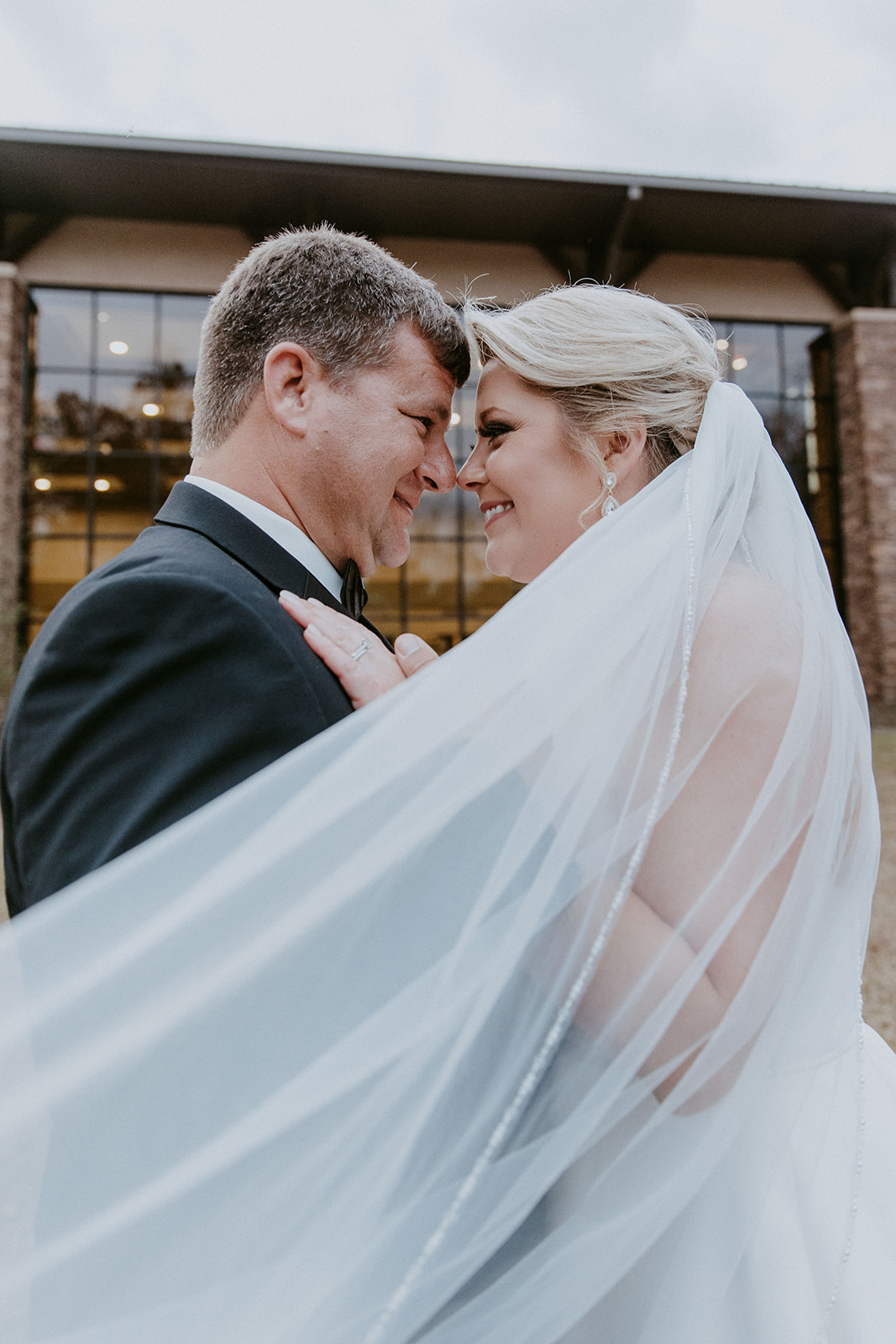 A bride and groom embrace outside a building with large windows. The bride's veil is flowing, and they both have their eyes closed. Trees and foliage are in the background at The Venues at Ogeechee Tech
