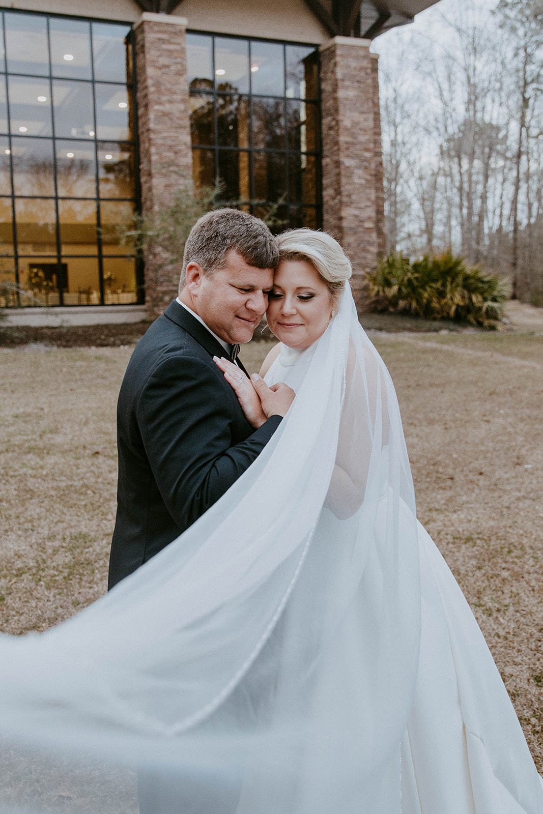 A bride and groom embrace outside a building with large windows. The bride's veil is flowing, and they both have their eyes closed. Trees and foliage are in the background at The Venues at Ogeechee Tech
