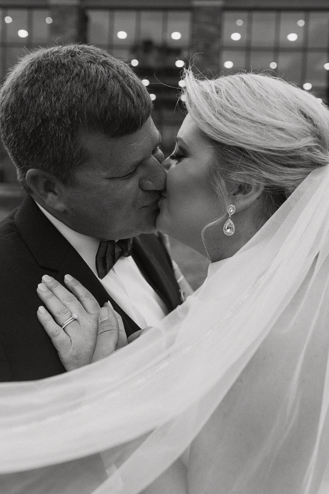 A bride and groom embrace outside a building with large windows. The bride's veil is flowing, and they both have their eyes closed. Trees and foliage are in the background at The Venues at Ogeechee Tech
