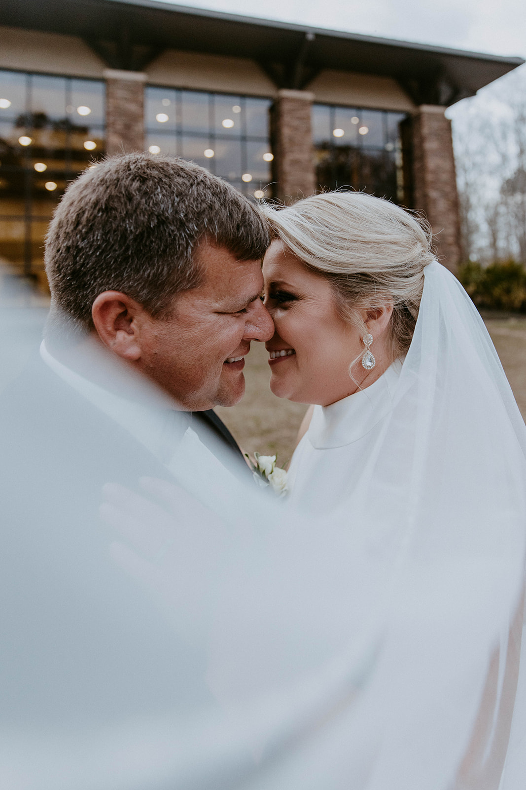 A bride and groom embrace outside a building with large windows. The bride's veil is flowing, and they both have their eyes closed. Trees and foliage are in the background at The Venues at Ogeechee Tech
