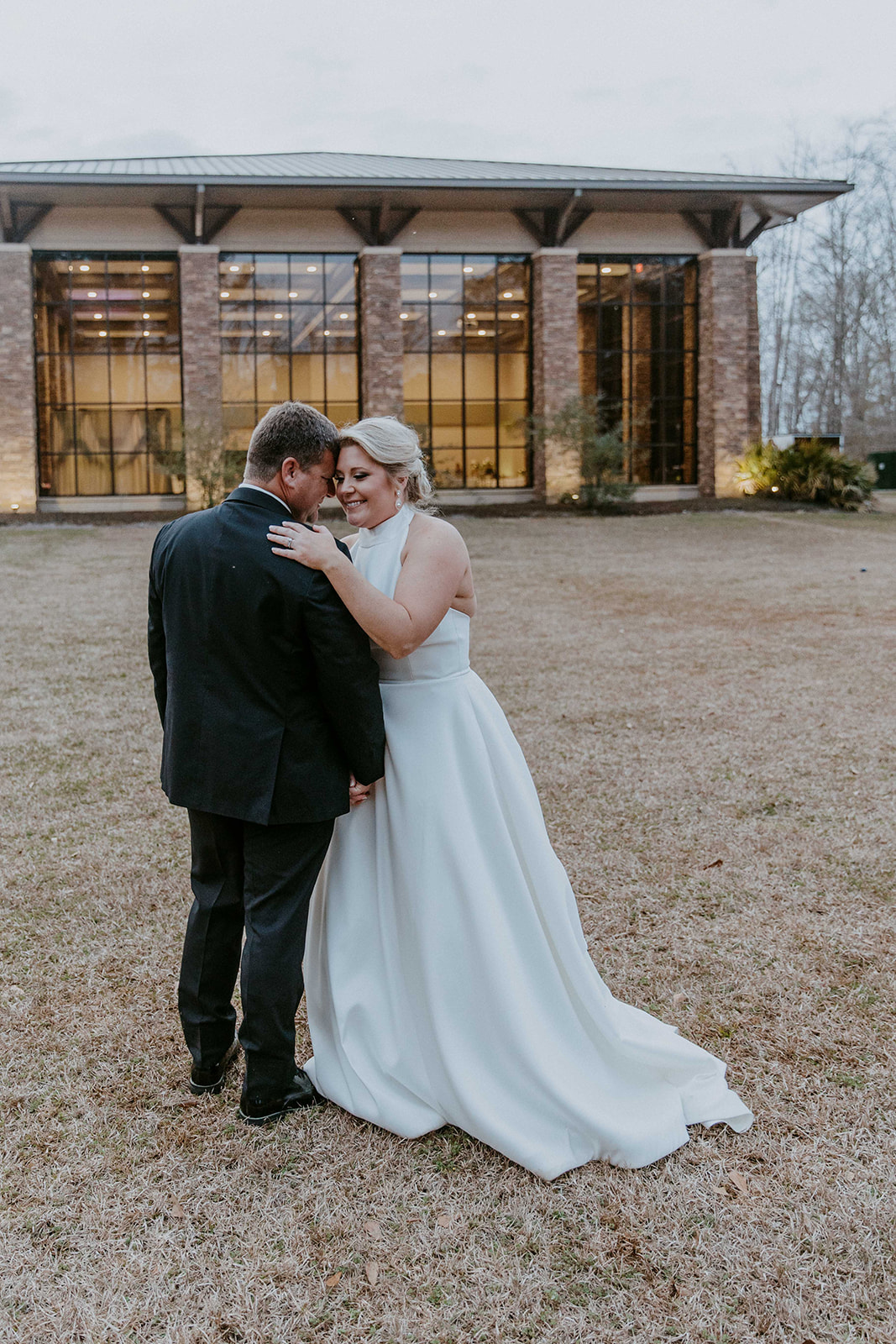 A bride and groom embrace outside a building with large windows. The bride's veil is flowing, and they both have their eyes closed. Trees and foliage are in the background at The Venues at Ogeechee Tech
