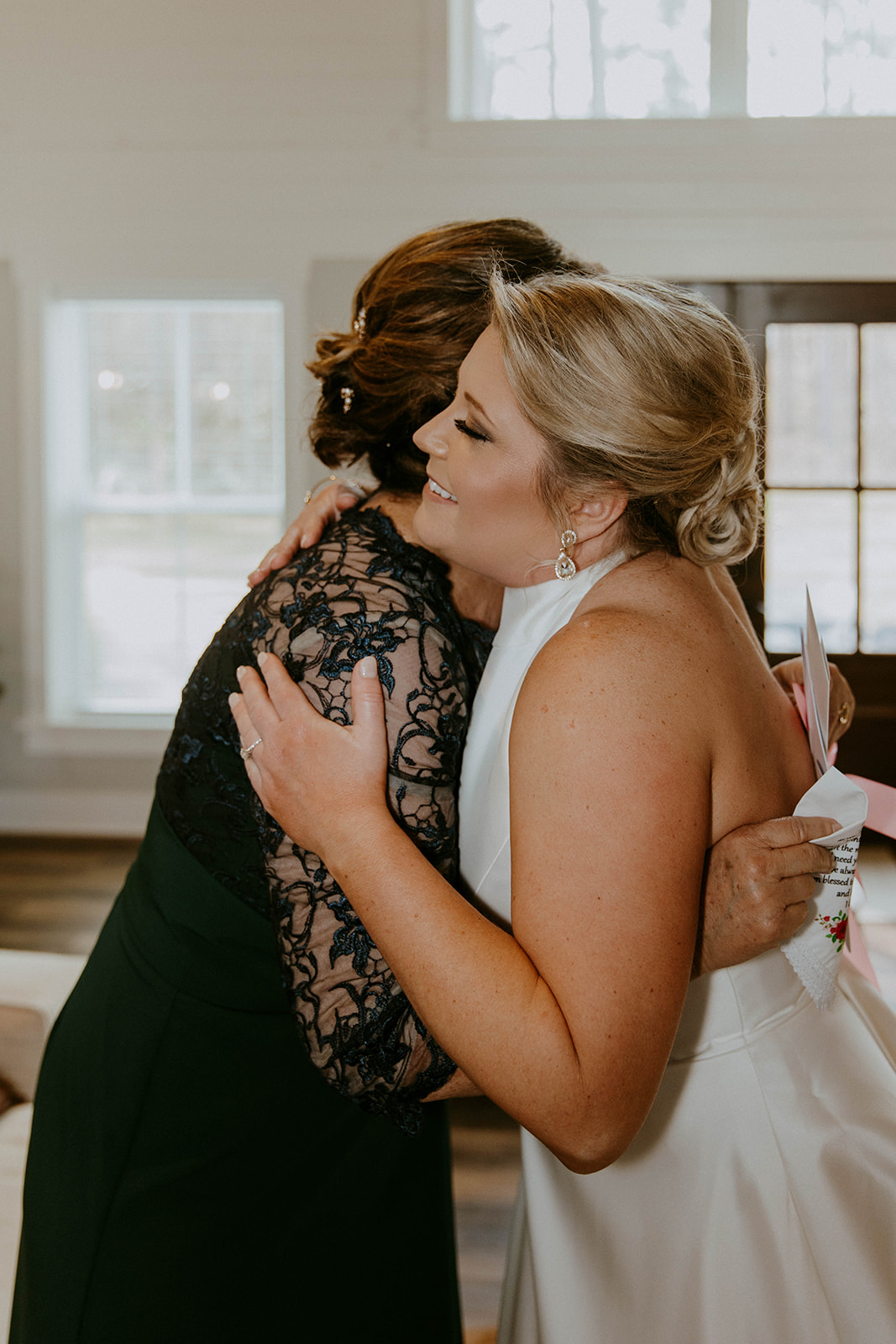 A bride is being assisted by two women as they adjust the back of her white wedding dress. They are indoors, and the room has wood flooring and natural light from the windows at The Venues at Ogeechee Tech