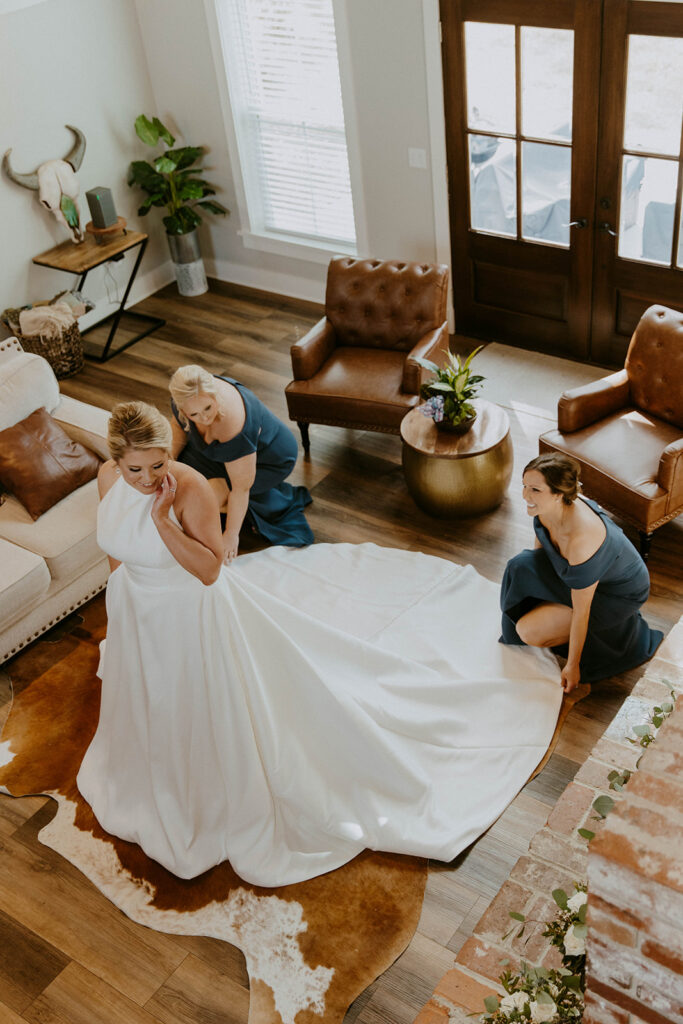 A bride is being assisted by two women as they adjust the back of her white wedding dress. They are indoors, and the room has wood flooring and natural light from the windows at The Venues at Ogeechee Tech