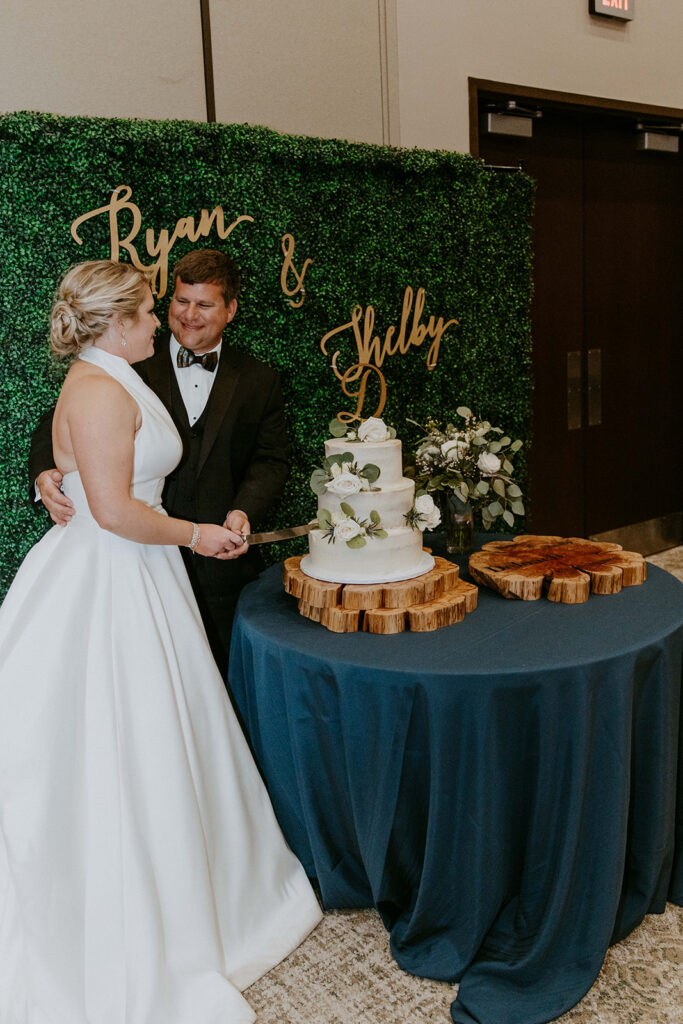 A bride and groom stand in front of a green wall with "Ryan & Shelby" written on it, cutting a two-tiered white wedding cake on a blue tablecloth.