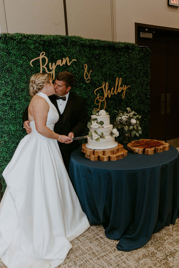 A bride and groom stand in front of a green wall with "Ryan & Shelby" written on it, cutting a two-tiered white wedding cake on a blue tablecloth.