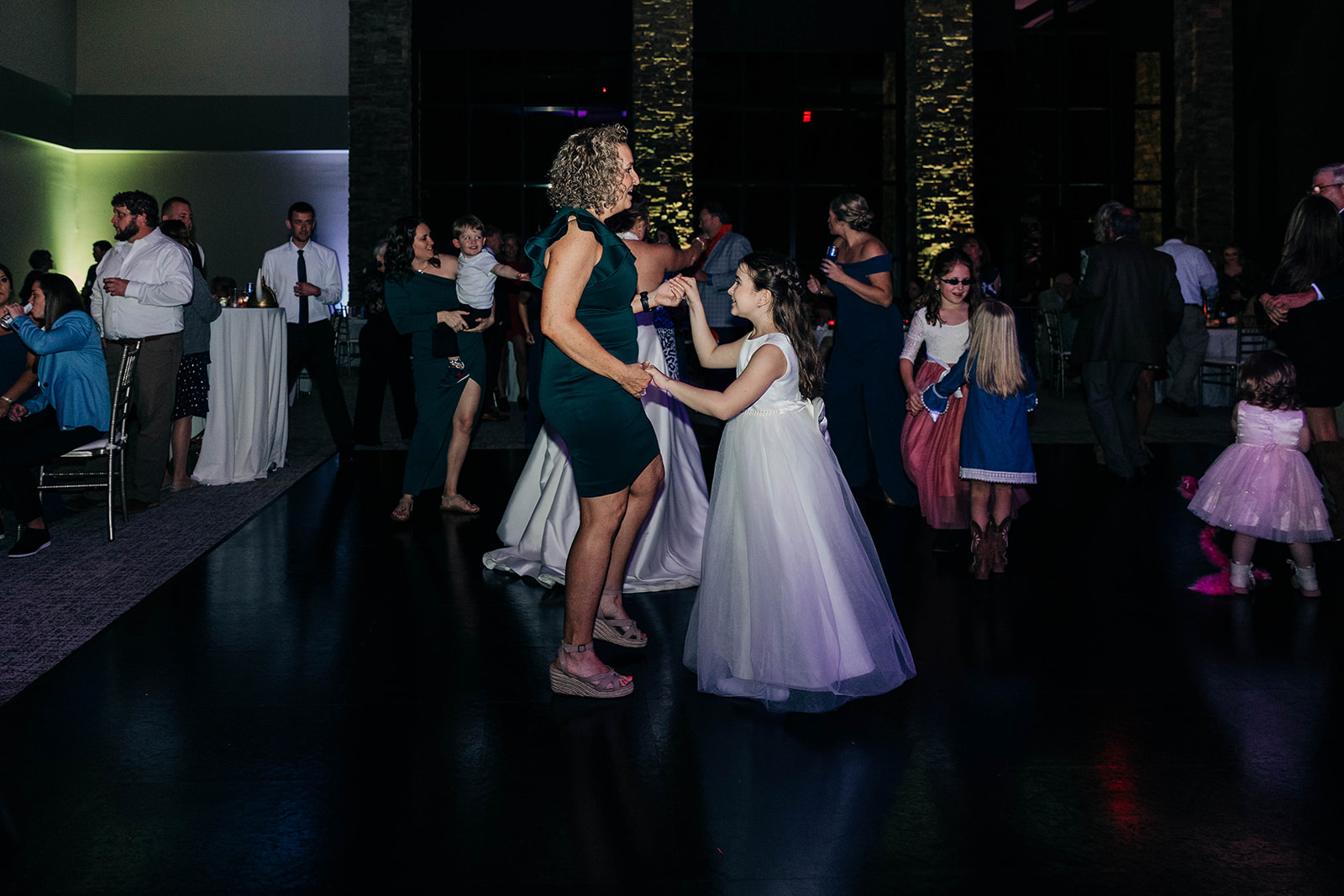 People dressed formally dance in a dimly lit room at what appears to be a wedding reception. A woman in a green dress dances with a young girl in a white dress in the center of the image.