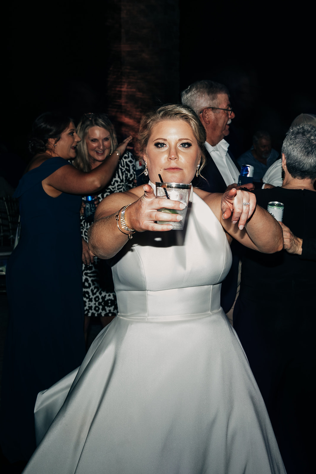 People dressed formally dance in a dimly lit room at what appears to be a wedding reception. A woman in a green dress dances with a young girl in a white dress in the center of the image.