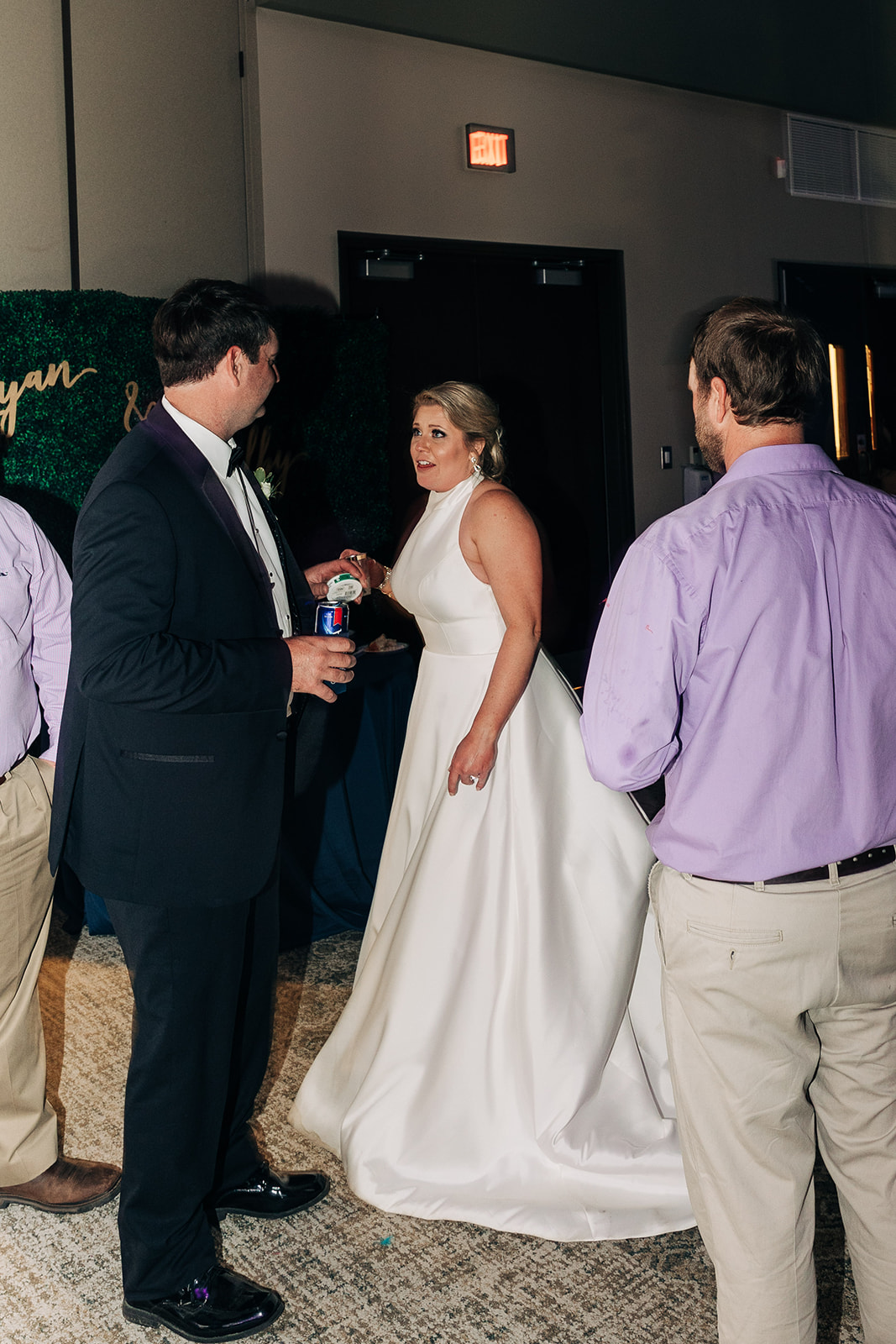 People dressed formally dance in a dimly lit room at what appears to be a wedding reception. A woman in a green dress dances with a young girl in a white dress in the center of the image.