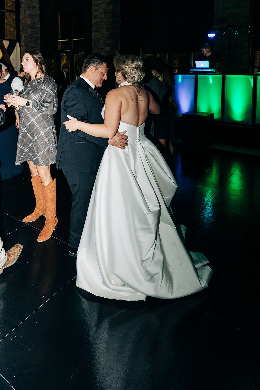 People dressed formally dance in a dimly lit room at what appears to be a wedding reception. A woman in a green dress dances with a young girl in a white dress in the center of the image.