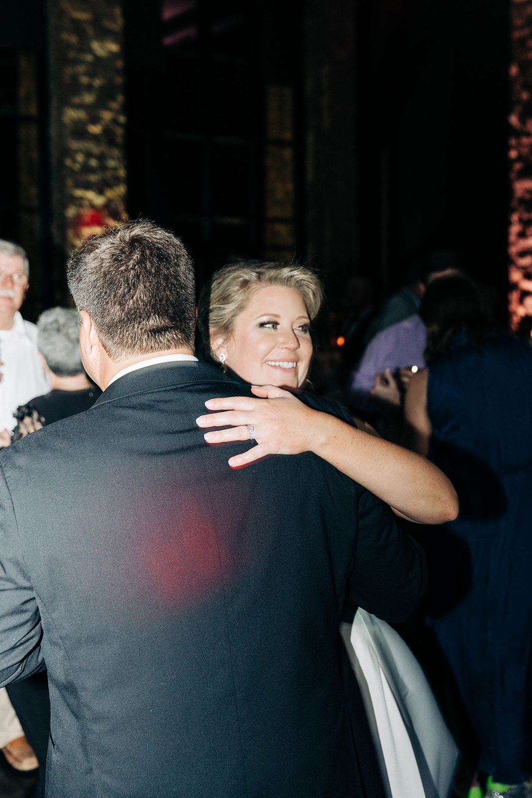 People dressed formally dance in a dimly lit room at what appears to be a wedding reception. A woman in a green dress dances with a young girl in a white dress in the center of the image.