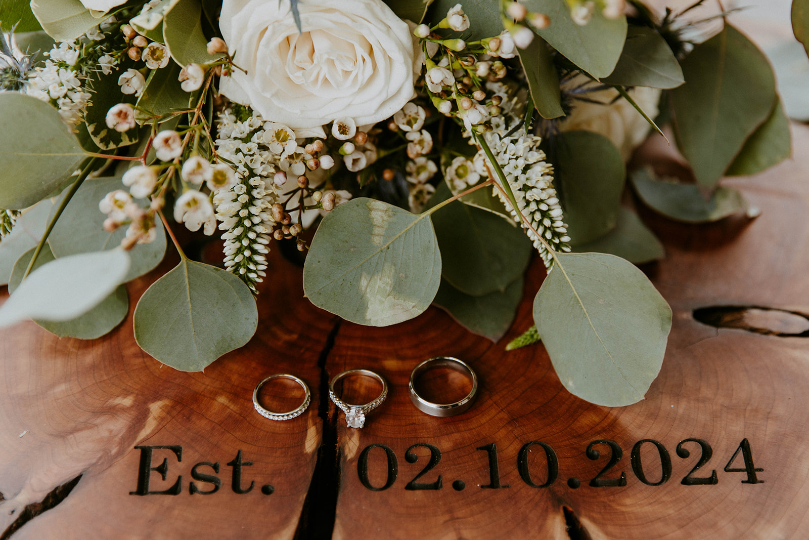 Close-up of three rings on a wooden surface with a bouquet of white flowers and green foliage. The text "Est. 02.10.2024" is printed below the rings at a wedding at the venues at ogeechee tech