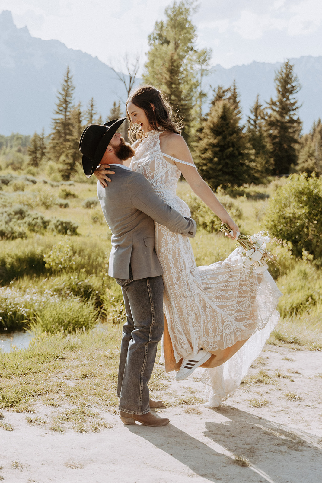 A man in a cowboy hat lifts a woman in a white dress in a grassy area with trees and mountains in the background- must have wedding poses