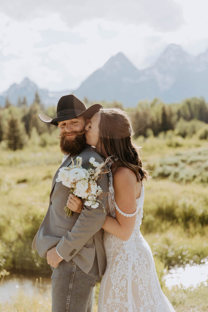 A bride and groom, dressed casually with flourishes of traditional wedding attire, share a kiss in a scenic outdoor setting with mountains and greenery in the background.