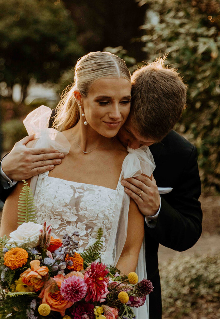 A bride and groom, dressed casually with flourishes of traditional wedding attire, share a kiss in a scenic outdoor setting with mountains and greenery in the background.