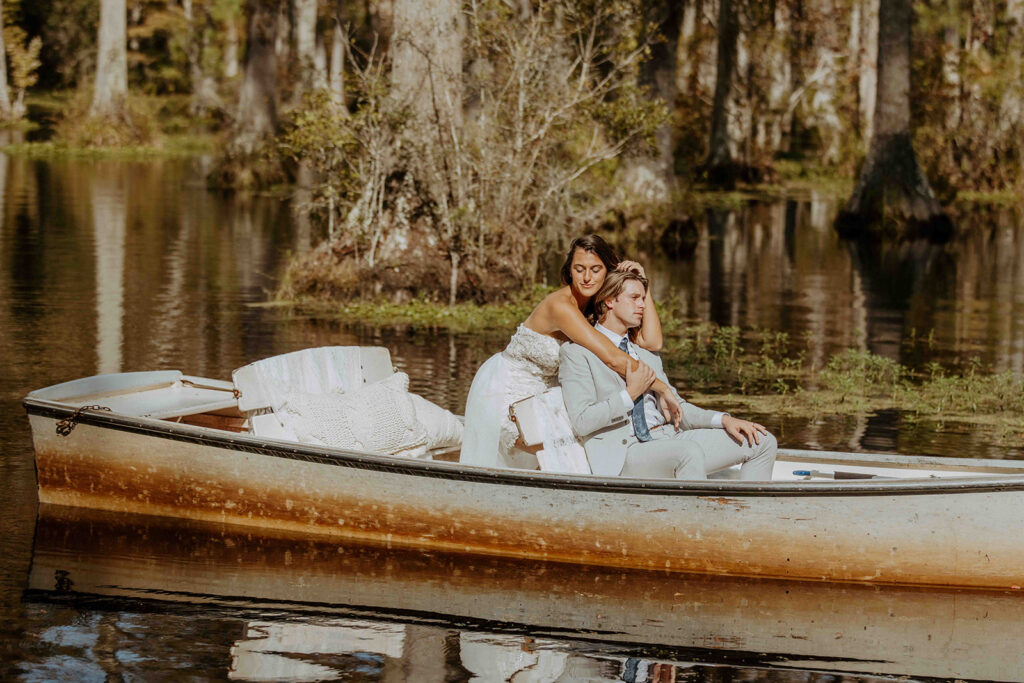 Two women in casual clothes sit together on a small boat in a calm, forested lake, surrounded by trees and greenery.