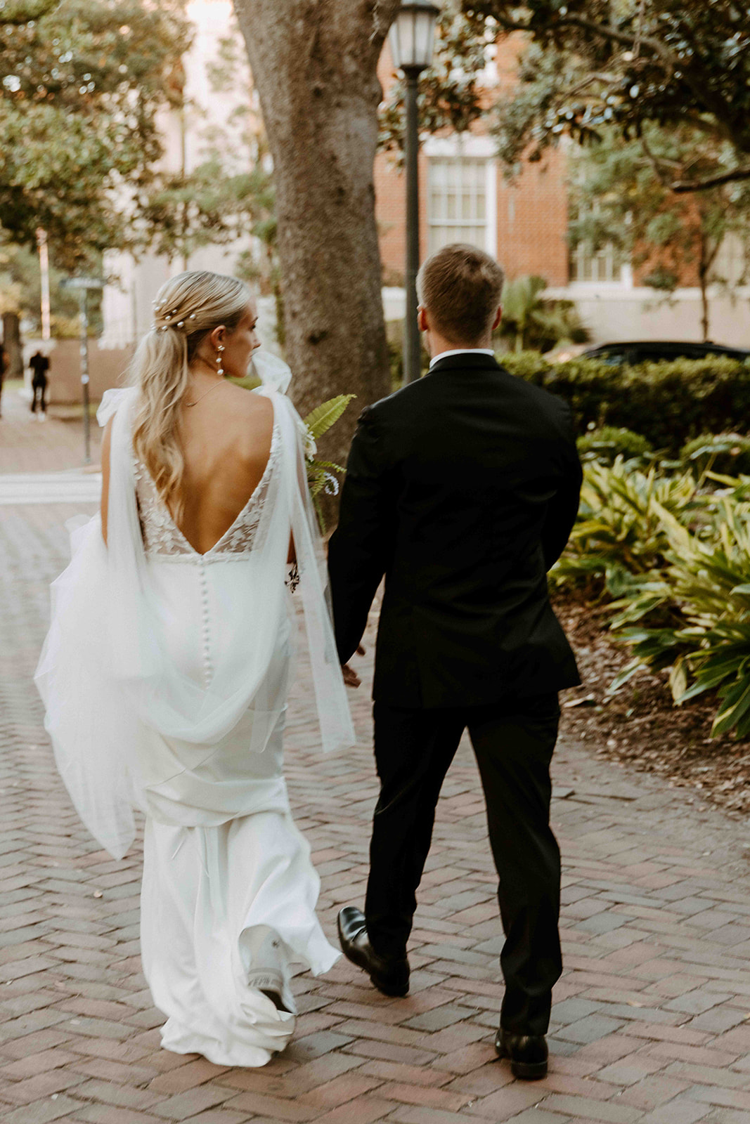 A couple, seen from behind, wearing formal attire walks hand in hand along a brick path. The woman is in a white dress with a low back, and the man is in a black suit. Trees and greenery surround them- must have wedding poses