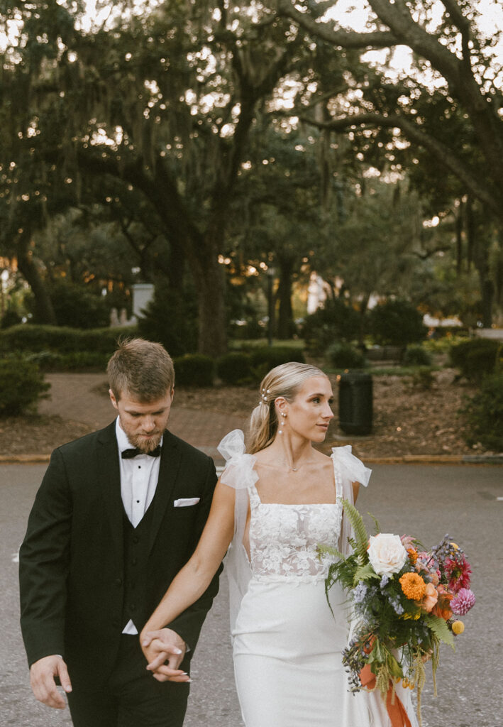 A couple in formal attire, holding hands and walking outdoors; the man in a black tuxedo and the woman in a white wedding dress holding a colorful bouquet. Trees and greenery are visible in the background.