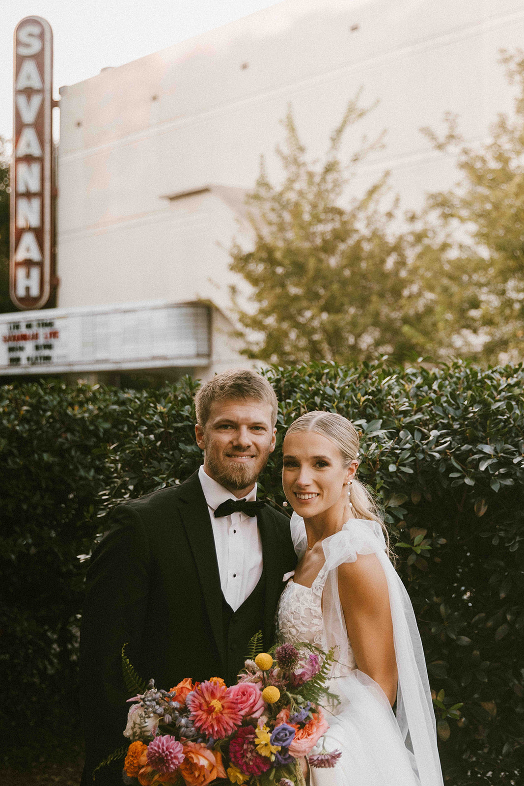 A couple dressed in wedding attire stands close together, smiling. The bride holds a colorful bouquet. The background shows greenery and a building with a "Savannah" sign. -must have wedding poses