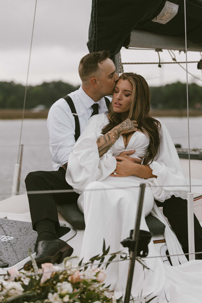 A bride and groom, dressed casually with flourishes of traditional wedding attire, share a kiss in a scenic outdoor setting with mountains and greenery in the background.