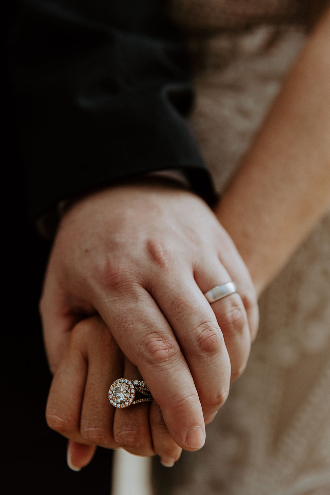 A couple stands together, the woman displaying an engagement ring on her hand as she looks up at the man who has his arms wrapped around her.