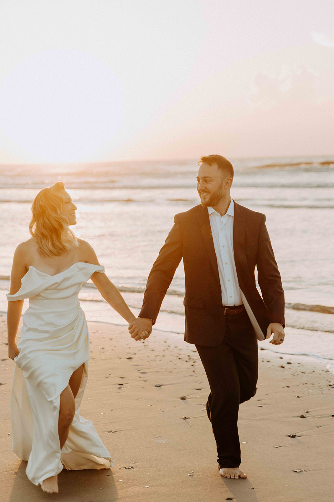 A couple, dressed in formal attire, walk barefoot on a beach at sunset. The woman wears a white dress, and the man is in a dark suit -must have wedding poses