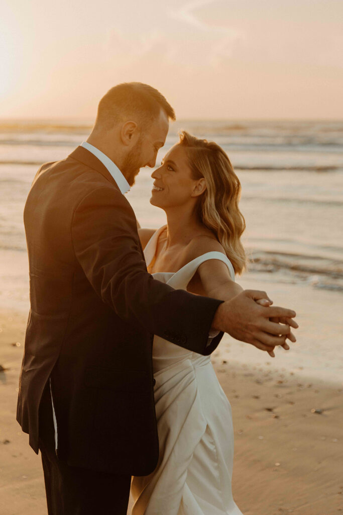 A couple dressed in formal attire dances on a beach at sunset, looking at each other with smiles.