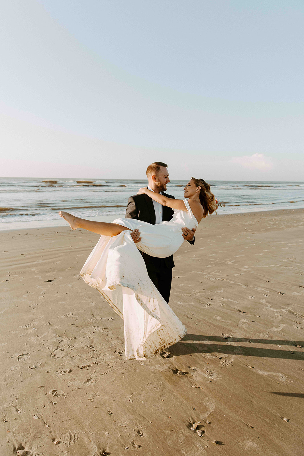 A man in a suit holds a woman in a white dress in his arms on a sandy beach with waves in the background- must have wedding poses